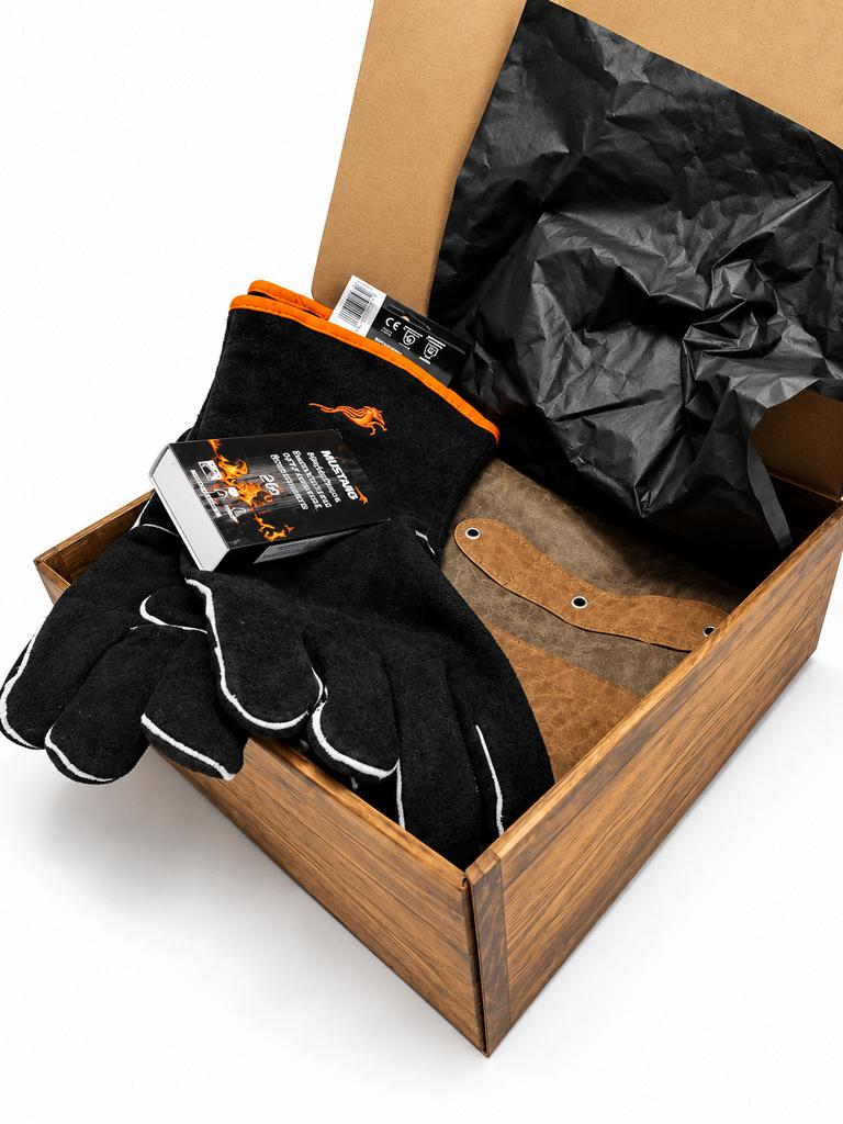 Geöffnete Grill-Geschenkbox mit Mustang Leder-Grillhandschuhen, brauner Grillschürze und BBQ-Streichhölzern auf schwarzem Geschenkpapier